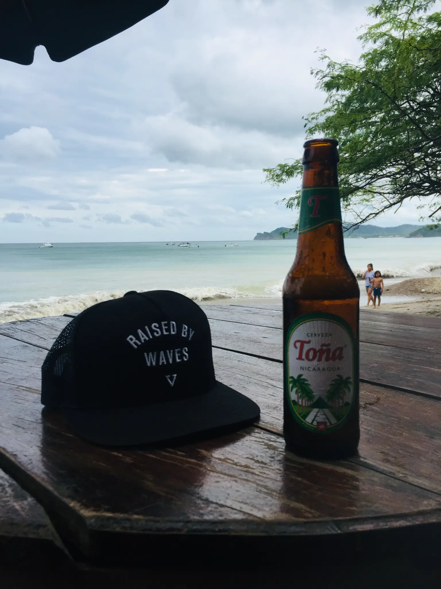 Raised by Waves hat and cold Toña Nicaragua beer on a wooden table with the emerald Pacific beach behind