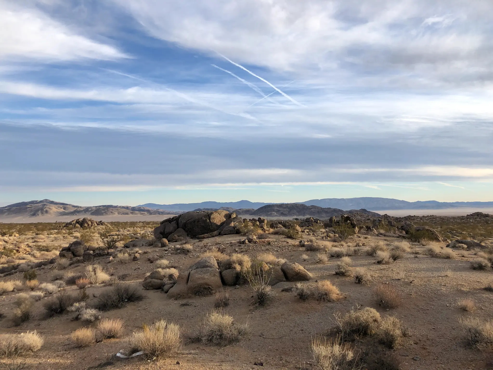 Wide Mojave desert panorama near Landers, CA — granite boulders, desert scrub, and mountain ranges under a dramatic cloud-streaked sky