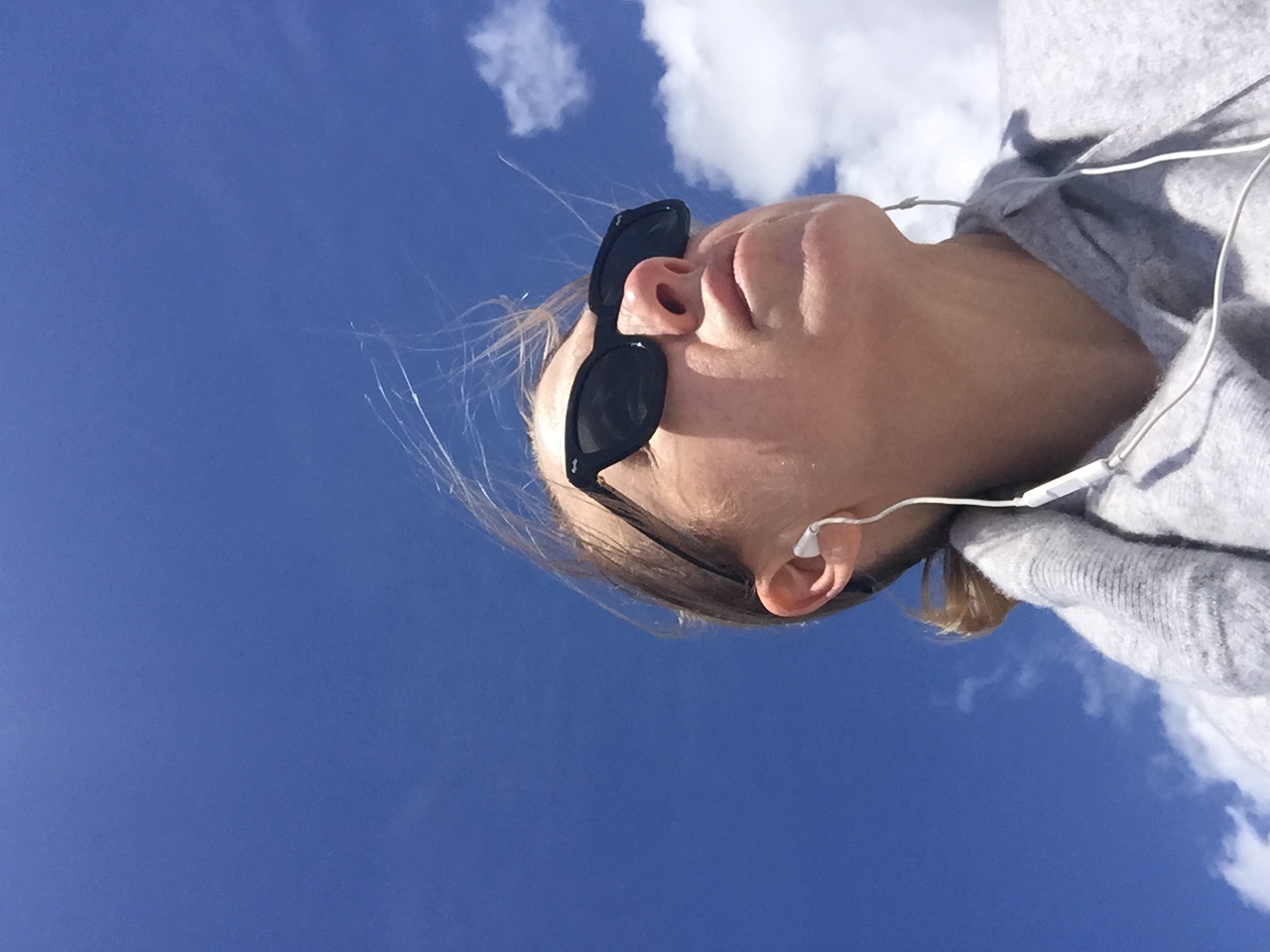 Kristen looking up at a bright blue sky with earbuds in, wind in her hair — taken during a walk while waiting